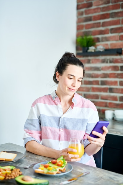 Taille hoch Porträt einer lächelnden jungen Frau, die das Frühstück zu Hause genießt und soziale Medien überprüft