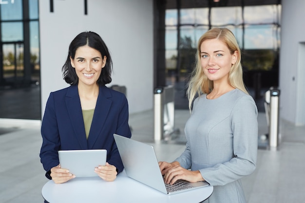 Taille hoch Porträt von zwei erfolgreichen Geschäftsfrauen, die an der Kamera lächeln, während am Schreibtisch im Bürogebäude stehen und Laptop halten,