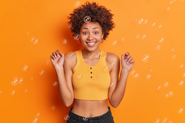 Taille hoch Schuss der fröhlichen afroamerikanischen Frau hebt die Hände lächelt breit trägt kurz geschnittenes Oberteil und Jeans hat perfekte Figur drückt positive Emotionen aus, die über orangefarbenen Wandblasen isoliert sind