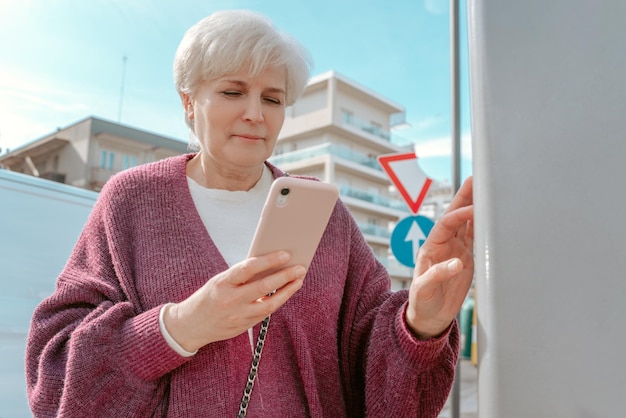 Taille-Up-Porträt einer ernsten, konzentrierten, grauhaarigen Dame, die auf ihr Smartphone schaut, während sie neben dem Auto steht