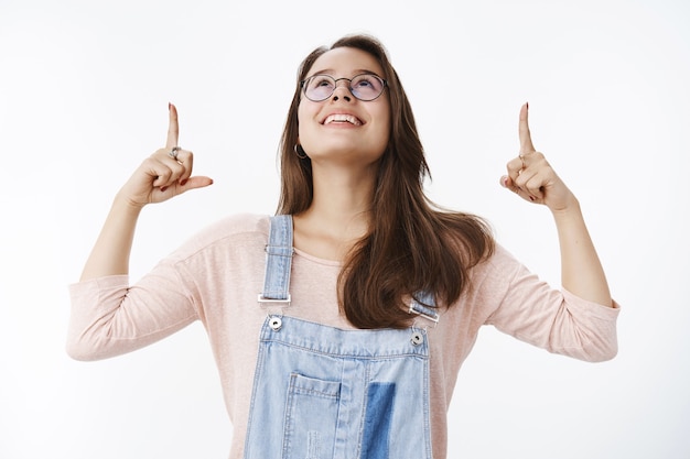 Taillenaufnahme einer amüsierten glücklichen europäischen Frau in Brille und Overall, die den Kopf hebt und nach oben zeigt, fasziniert von einem faszinierten und erfreuten Lächeln, das gegen eine graue Wand posiert. Platz kopieren