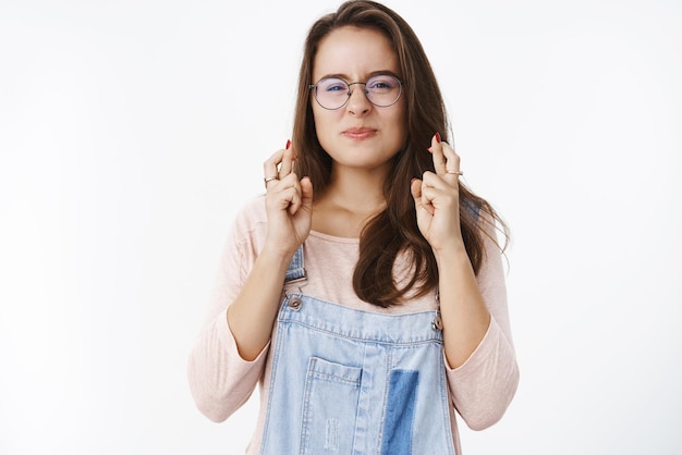 Taillenaufnahme einer hoffnungsvollen, glücklichen und optimistischen süßen Frau mit Brille, die die Nase schielt und kräuselt, als eifrig zu wünschen, dass die Daumen wahr werden, um viel Glück beim Träumen und Beten aufgeregt über der grauen Wand zu bekommen