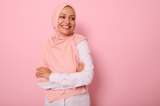 Taillenlanges Porträt einer schönen arabisch-muslimischen Frau in rosa Hijab, die mit attraktivem Blick, zahnigem Lächeln, selbstbewusstem Aussehen und verschränkten Armen zur Seite schaut, auf rosafarbenem Hintergrund mit Kopierraum