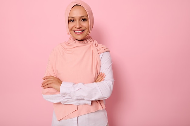 Taillenlanges Porträt einer wunderschönen arabisch-muslimischen Frau in rosa Hijab, die mit attraktivem Blick, zahnigem Lächeln, selbstbewusstem Blick und verschränkten Armen auf die Kamera blickt, auf rosafarbenem Hintergrund mit Kopierraum