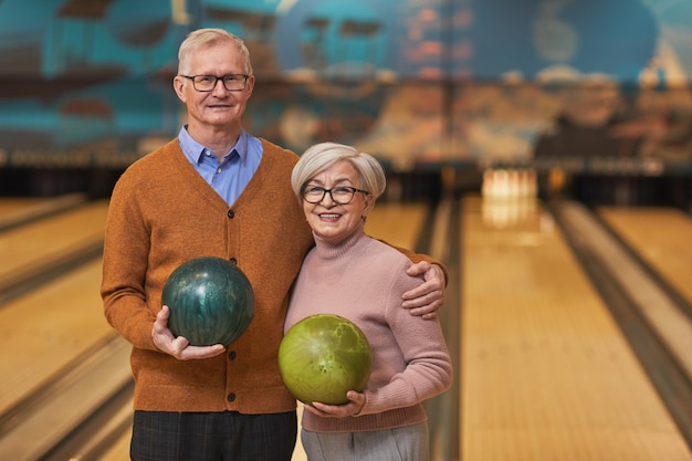 Taillenporträt eines glücklichen Seniorenpaares, das Bowlingkugeln hält und in die Kamera lächelt, während Sie aktive Unterhaltung auf der Bowlingbahn genießen, Platz kopieren