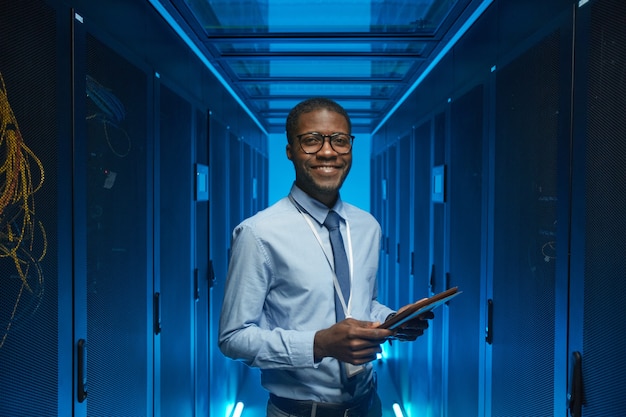 Taillenporträt eines lächelnden afroamerikanischen Mannes, der am Serverschrank steht, während er mit einem Supercomputer im Rechenzentrum arbeitet und Tablet hält, Platz kopieren