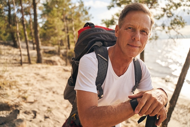 Taillenporträt eines lächelnden sportlichen Mannes mit Rucksack beim Gehen mit Wanderstöcken im Wald in der Nähe des Meeres