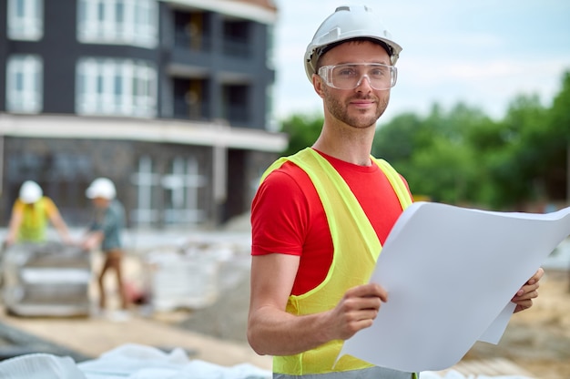 Taillenporträt eines lächelnden süßen jungen Mannes mit Hausplänen, der für die Kamera auf der Baustelle posiert