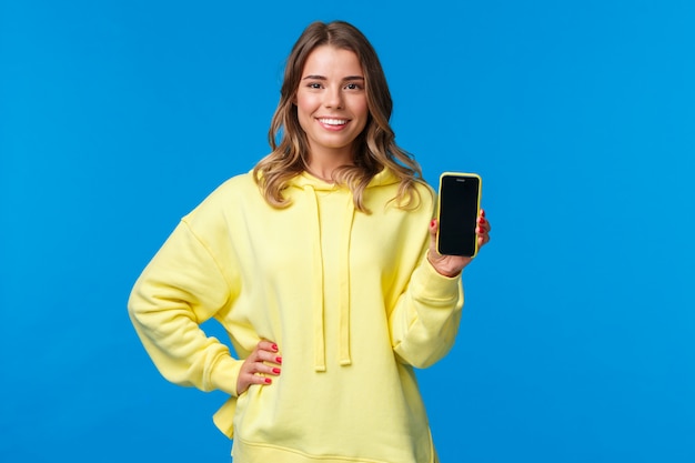 Taillenporträt gut aussehendes blondes Mädchen im gelben Kapuzenpulli fördern Smartphone-Anwendung, zeigen Handy-Anzeige und lächeln als empfehlen abonnieren oder App herunterladen