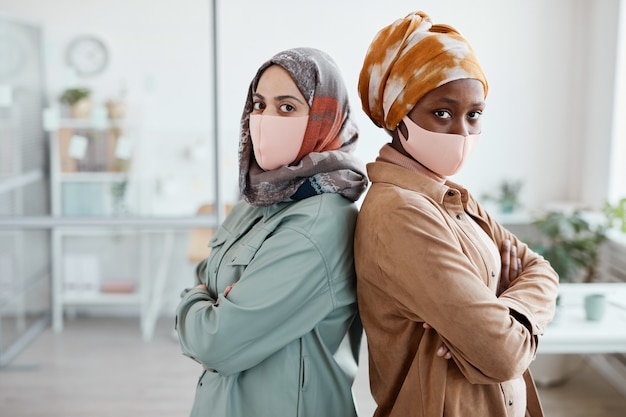 Taillenporträt von zwei ethnischen jungen Frauen, die Rücken an Rücken im Büro stehen und in die Kamera schauen, Platz kopieren