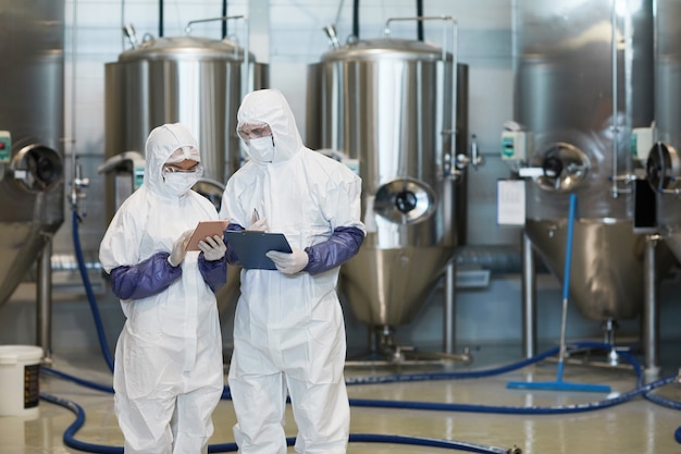 Taillenporträt von zwei jungen Arbeitern, die Hazmat-Anzüge tragen, während sie ein digitales Tablet in einer modernen Chemiefabrik verwenden, Platz kopieren