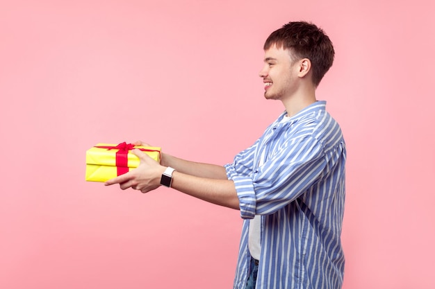 Take it Porträt eines positiven, großzügigen braunhaarigen Mannes mit kleinem Bart im Freizeithemd, der der linken Seite ein Geschenk gibt und zum Geburtstag gratuliert, Studioaufnahme isoliert auf rosa Hintergrund