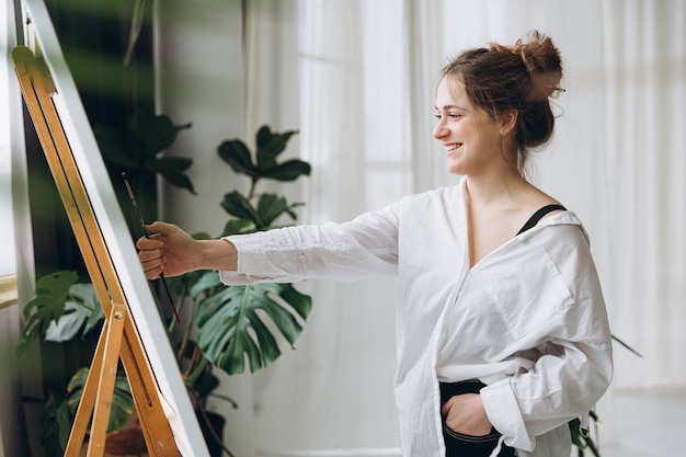 Talentierte Künstlerin in lässigem Outfit mit Pinsel zum Messen auf Leinwand