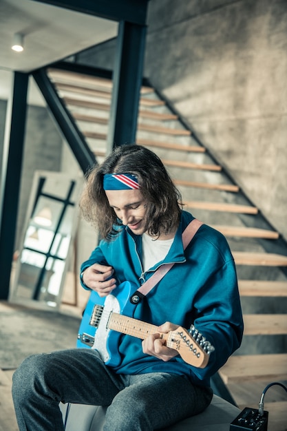 Foto talento exclusivo. sonriente chico de pelo largo siendo feliz mientras toca la guitarra electrónica en sus elegantes apartamentos