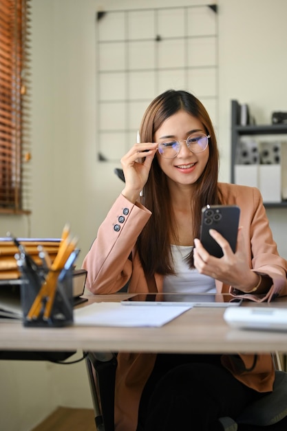 Una talentosa mujer de negocios asiática con anteojos usando su teléfono móvil en el escritorio de su oficina