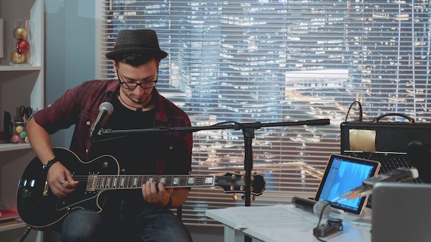 Talentoso guitarrista con sombrero y ropa casual de moda tocando la guitarra y cantando en el estudio de grabación en casa