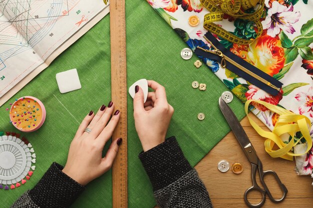 Taller de costura o diseñador de moda en el trabajo. Vista superior de las manos femeninas que marcan el cuadrado de tela con un trozo de jabón y una regla. Mesa desordenada con accesorios y patrones de modista, espacio de copia