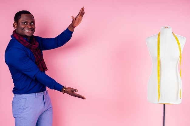 Taller de costurera de sastre de hombre afroamericano, diseñador de ropa de modelo masculino elegante posando junto al maniquí sobre fondo rosa en el estudio, las manos apuntan al espacio de diseño copyspace