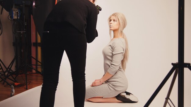 Taller detrás del escenario - chica guapa rubia posando para el fotógrafo - modelo se sienta en las rodillas, fondo blanco.