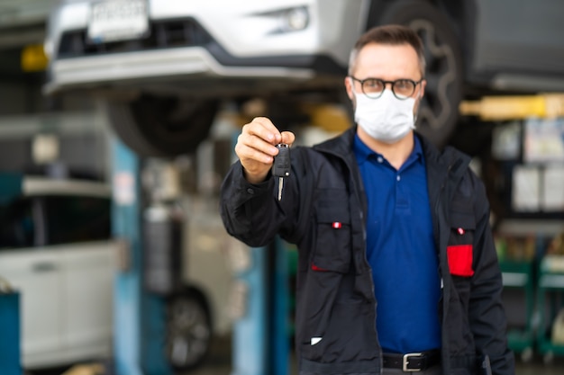 Taller de mantenimiento y servicio de automóviles. El gerente de Portrait Beard Male da una llave del auto a la cámara y usa mascarilla médica de protección contra el coronavirus.