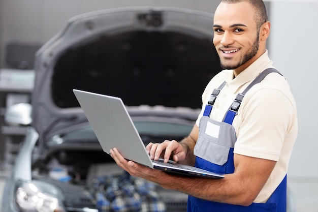 Foto taller de reparación de automóviles, mecánico masculino cerca del automóvil con computadora portátil