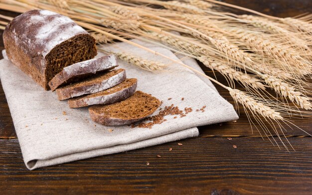 Tallos de tallos y granos de trigo integral secos junto a una sola barra de delicioso pan de centeno con rebanadas de tela marrón sobre la mesa de madera oscura