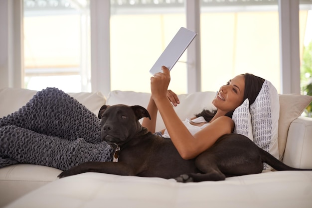También tienen aplicaciones para mascotas en estos días Foto de una mujer joven y atractiva relajándose en el sofá con su perro y usando una tableta digital