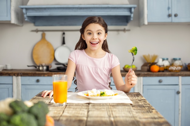 Tan delicioso. Hermoso contenido niña morena sonriendo y desayunando saludablemente y bebiendo jugo