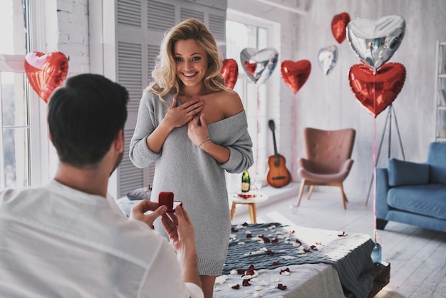 ¡Tan feliz! Mujer joven sorprendida mirando el anillo de compromiso con una sonrisa mientras su novio le propone en el dormitorio