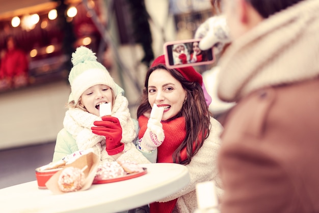 Tan hambriento. Hembras encantadas comiendo dulces mientras pasan tiempo juntos en el mercado navideño