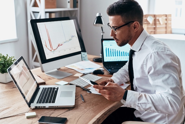 Foto tan ocupado. joven empresario en ropa formal usando un teléfono inteligente y analizando datos usando la computadora mientras está sentado en la oficina