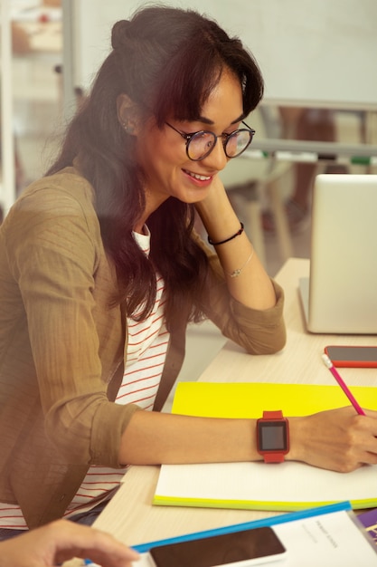 Tan tímido. Bastante joven mujer manteniendo una sonrisa en su rostro mientras estudia con placer