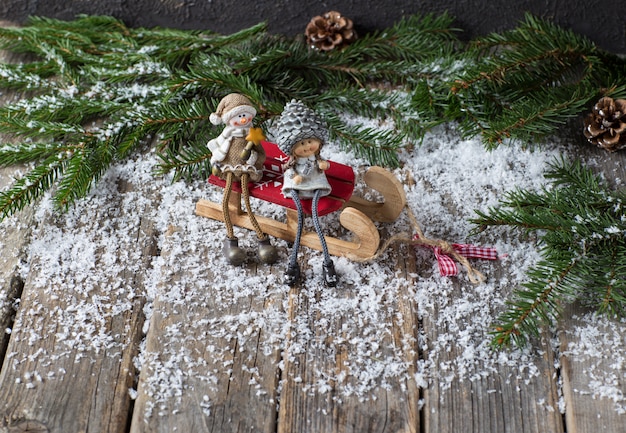 Tannenzweige, Zapfen, Schnee und zwei Figuren (ein Schneemann und ein Mädchen) sitzen auf einem Holztisch