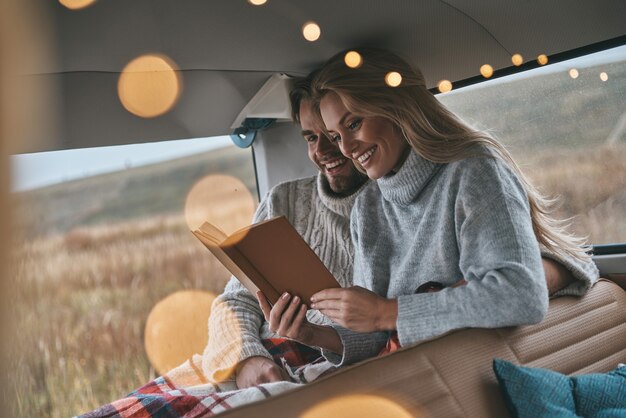 Tão interessante. Lindo casal jovem lendo o livro e sorrindo enquanto está sentado em uma minivan estilo retro