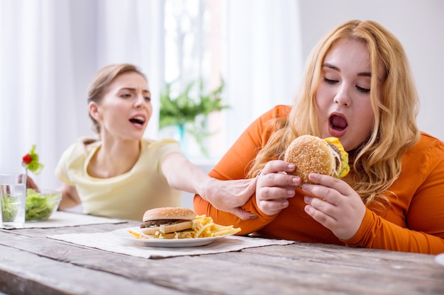 Tão saboroso. Mulher gorda e obstinada comendo um sanduíche e sua amiga a impedindo