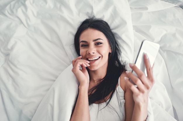 Tão satisfeito. Menina morena encantada deitada em sua cama e segurando o telefone na mão enquanto conversa online