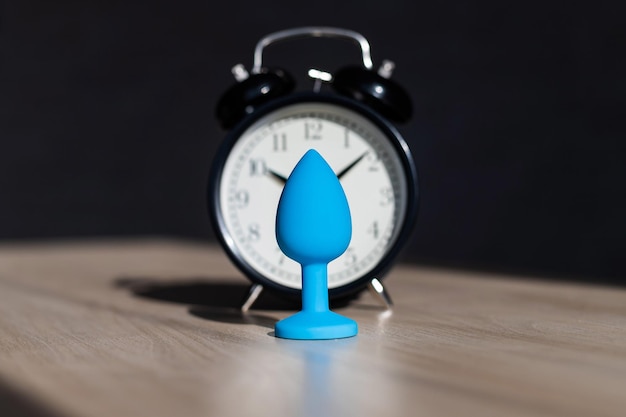 Tapón anal de látex azul y despertador sobre una mesa de madera La hora de los placeres carnales