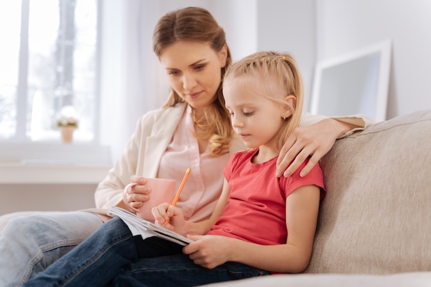 Tarea escolar. Grave chica inteligente agradable sentada junto con su madre y escribiendo en su cuaderno mientras hace una tarea en casa