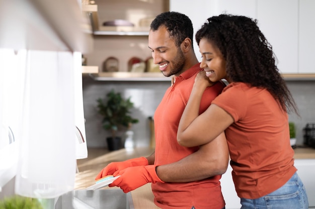 Tareas domésticas juntas en casa Una esposa negra milenaria feliz abrazando a su esposo con guantes de goma