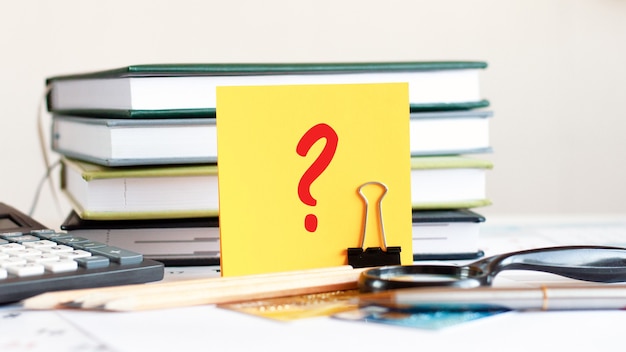 Una tarjeta amarilla con el signo de interrogación se encuentra en un clip para papeles en el escritorio con el telón de fondo de los libros, enfoque selectivo