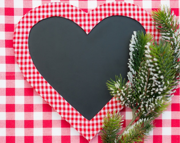 Tarjeta en blanco en forma de corazón con rama de árbol de Navidad sobre mantel a cuadros rojo Vacaciones de invierno