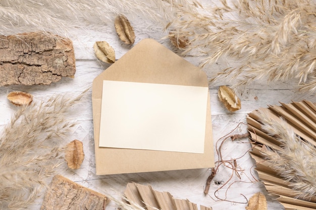 Tarjeta en blanco y sobre en la mesa de madera blanca cerca de plantas secas, hojas de palma y pasto de la pampa.