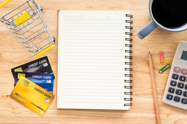 Tarjeta de crédito en carro de compras con cuaderno, lápiz, árbol de maceta, calculadora sobre fondo de madera, banca en línea Vista superior con mesa de oficina.