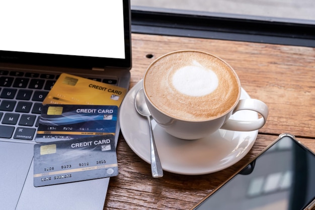 Tarjeta de crédito de computadora portátil, teléfono inteligente y café con leche caliente con espuma de leche latte art en taza sobre fondo de madera, concepto de banca en línea