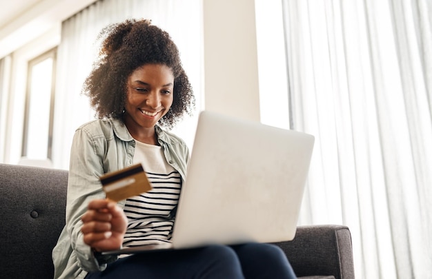 Tarjeta de crédito portátil y mujer africana en el sofá para transacción comercio electrónico o compras en línea en la sala de estar aplicación de pago y persona feliz para la compra suscripción o banca por Internet en el hogar