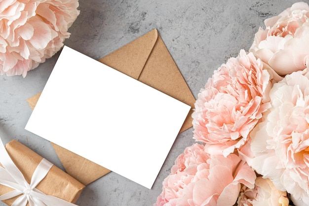 Tarjeta de felicitación en blanco en marco hecho de pastel color melocotón flores de peonía y caja de regalo en gris