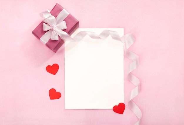 Tarjeta de felicitación del día de San Valentín con caja de regalo rosa, lazo blanco, cinta larga curvada y corazones rojos de papel. Vista superior, lugar para el texto