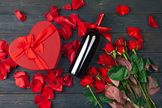 Tarjeta de felicitación del día de San Valentín. Flores rosas rojas, vino y caja de regalo en la mesa de superficie de madera.