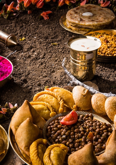 Tarjeta de felicitación Happy Holi diseñada mostrando comida dulce y salada tradicional india, flores y colores en polvo dispuestos sobre fondo blanco o arcilla. Enfoque selectivo