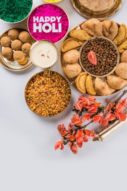 Tarjeta de felicitación Happy Holi diseñada mostrando comida dulce y salada tradicional india, flores y colores en polvo dispuestos sobre fondo blanco o arcilla. Enfoque selectivo